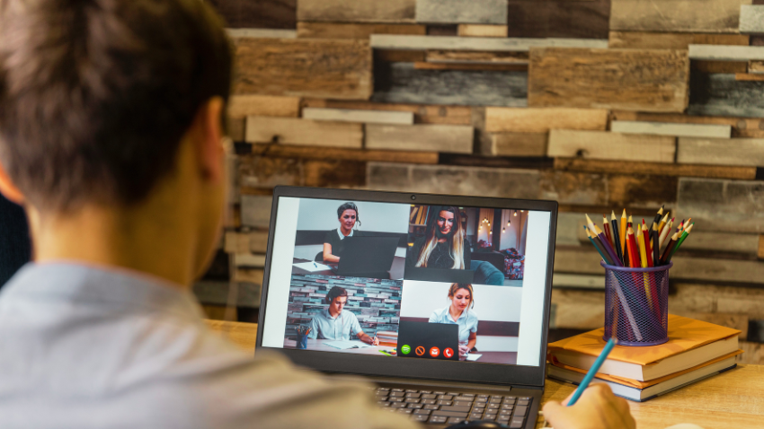 From a behind-the-shoulder perspective, a person with short brown hair is seen participating in a video conference call on a laptop. The laptop screen displays four participants in a grid layout. The person is holding a pencil over a notebook, suggesting they are taking notes. On the desk to the right, there's a purple mesh cup filled with colored pencils and a stack of books. The background features a textured wall of stacked stones or bricks.