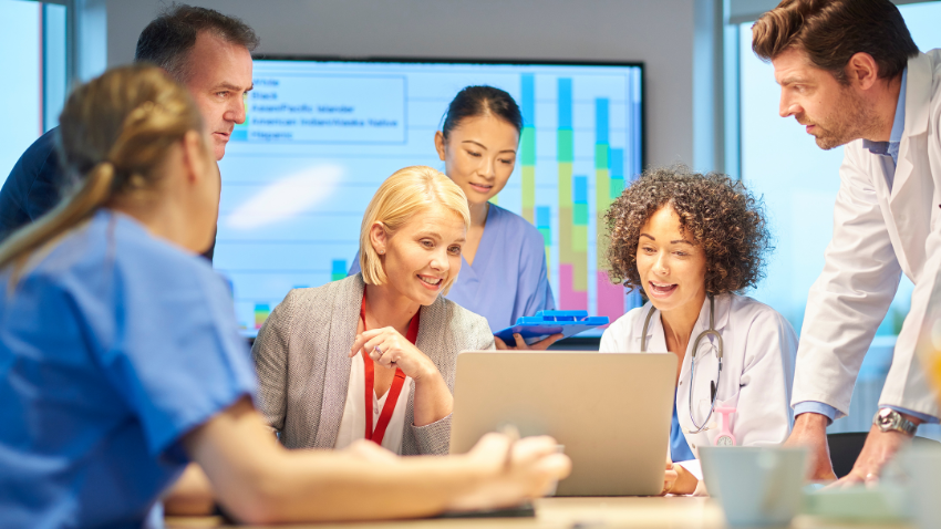 A diverse group of six healthcare professionals and administrators are gathered around a laptop in a modern meeting room. On the left, a man in a dark top and a person in blue scrubs look towards the center. In the middle, a blonde woman in a grey blazer and a woman in a purple scrub top are smiling and looking at the laptop screen. On the right, a woman in a white doctor's coat with a stethoscope around her neck is speaking and looking at the screen, while a man in a white lab coat stands over the group, looking on. A large screen in the background displays a bar chart.