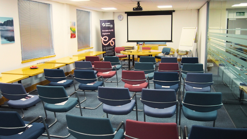 A brightly lit classroom or lecture hall with rows of empty chairs facing a projector screen at the front. The chairs are in various shades of blue and red. To the left, there are yellow desks along the wall under windows with blinds. In the front left corner, a banner with the "Open Awards" logo and "Access to HE Diplomas" is visible. A round table with chairs is in the center front, and a whiteboard easel is to the right of the screen. The room features a carpeted floor and glass partitions on the right.