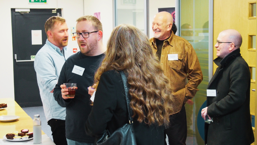 A group of five people are socializing in what appears to be an office or event space. On the left, two men are conversing, one holding a drink. In the center, a woman with long, curly brown hair, seen from behind, is talking to two bald men on the right. One of the bald men is smiling broadly, while the other is looking at the woman. There's a table with some snacks in the foreground on the left.
