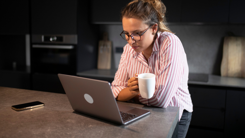 A woman with her hair pulled back and wearing glasses and a striped shirt leans on a dark countertop, looking intently at a silver laptop. She holds a white mug in her left hand. A smartphone lies on the counter to the left of the laptop. The background shows a dark kitchen with an oven and dark cabinets.