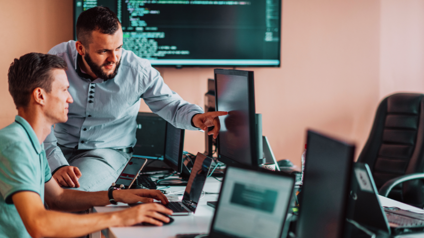 Two men are working together in an office setting. One man, wearing a light blue shirt, is seated at a desk looking at a laptop. Another man, with a beard and wearing a collared shirt, is leaning over the seated man's shoulder, pointing at the monitor. Several other computer screens and laptops are visible in the foreground and background, with code displayed on a large screen on the wall.