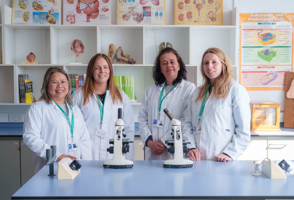 Four women, all wearing white lab coats and lanyards, are standing behind a lab bench with two microscopes on it, facing the camera and smiling. They are in what appears to be a science classroom or lab, with shelves behind them displaying anatomical charts, models, and specimens. A poster titled "Eukaryotic and Prokaryotic Cells" is visible on the far right.