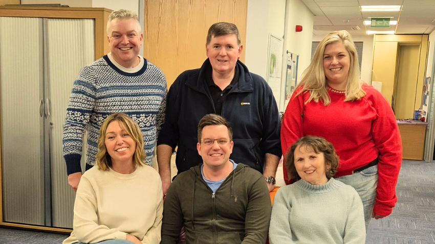A group of six smiling people are posing for a photo in an office setting. Three people are standing in the back row: a man on the left in a patterned blue and white sweater, a man in the middle in a dark blue fleece jacket, and a woman on the right in a bright red sweater. Three people are sitting in the front row: a woman on the left in a light sweater, a man in the middle in a green hoodie, and a woman on the right in a light blue sweater.