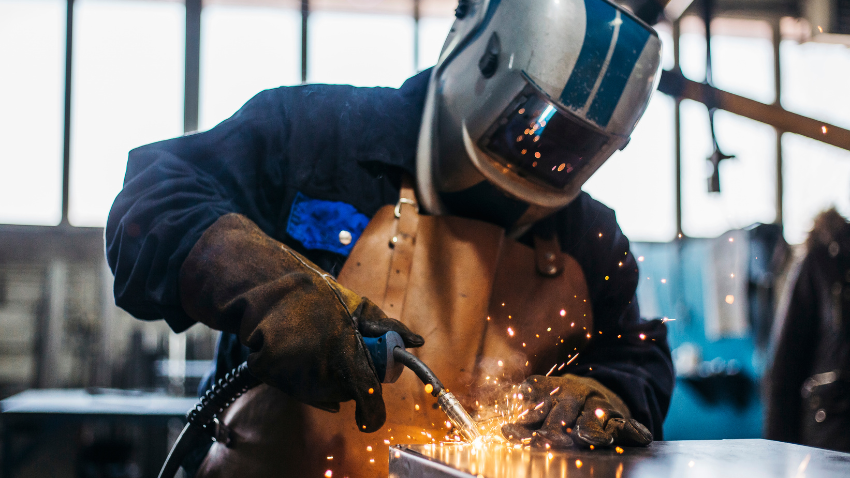 Welder wearing protective helmet and gloves performing metal welding in a workshop, with sparks flying during vocational skills training.